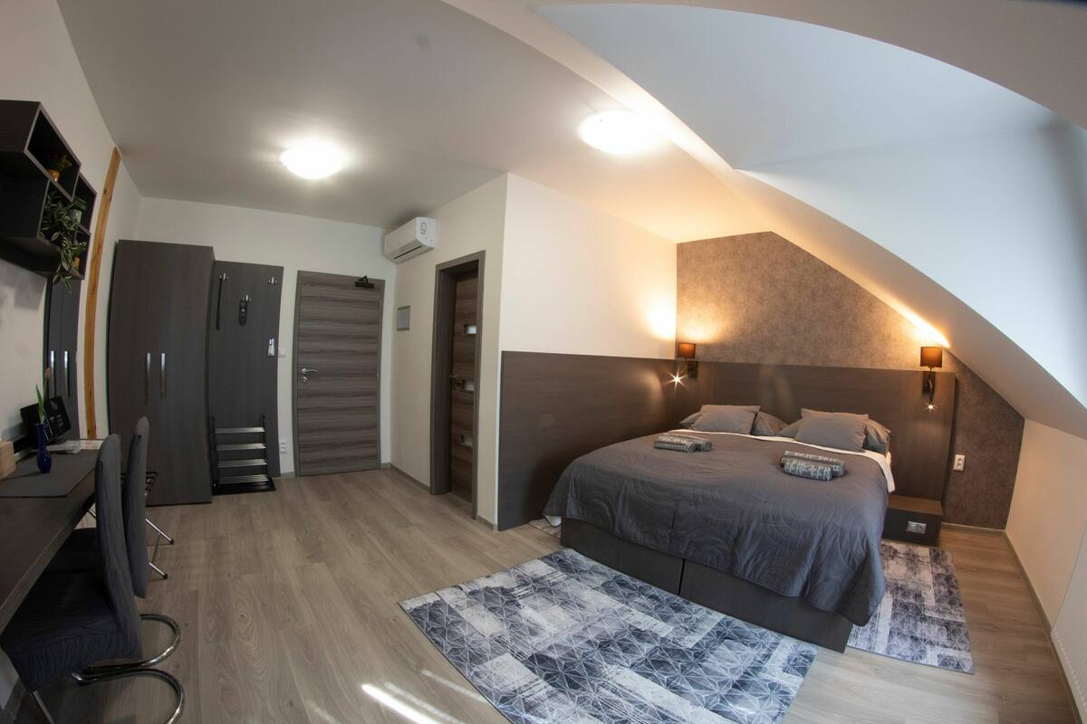 A modern bedroom with a sloped ceiling, a bed with a grey bedspread, a desk with a chair, and a wardrobe.