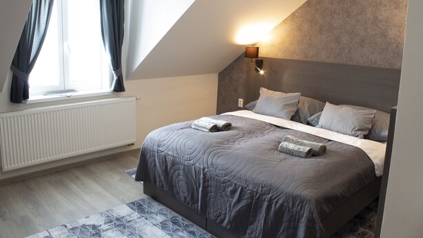 A modern bedroom with a sloped ceiling, a large bed with a grey quilt, a bedside table, and a wall-mounted light. - VALLA penzión&reštaurácia (Malacky)