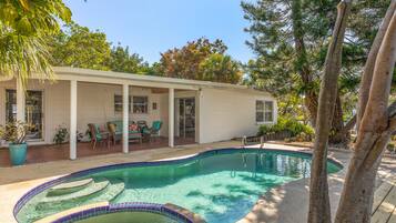 A swimming pool with a tiled edge, a covered patio with chairs, and a house in the background.