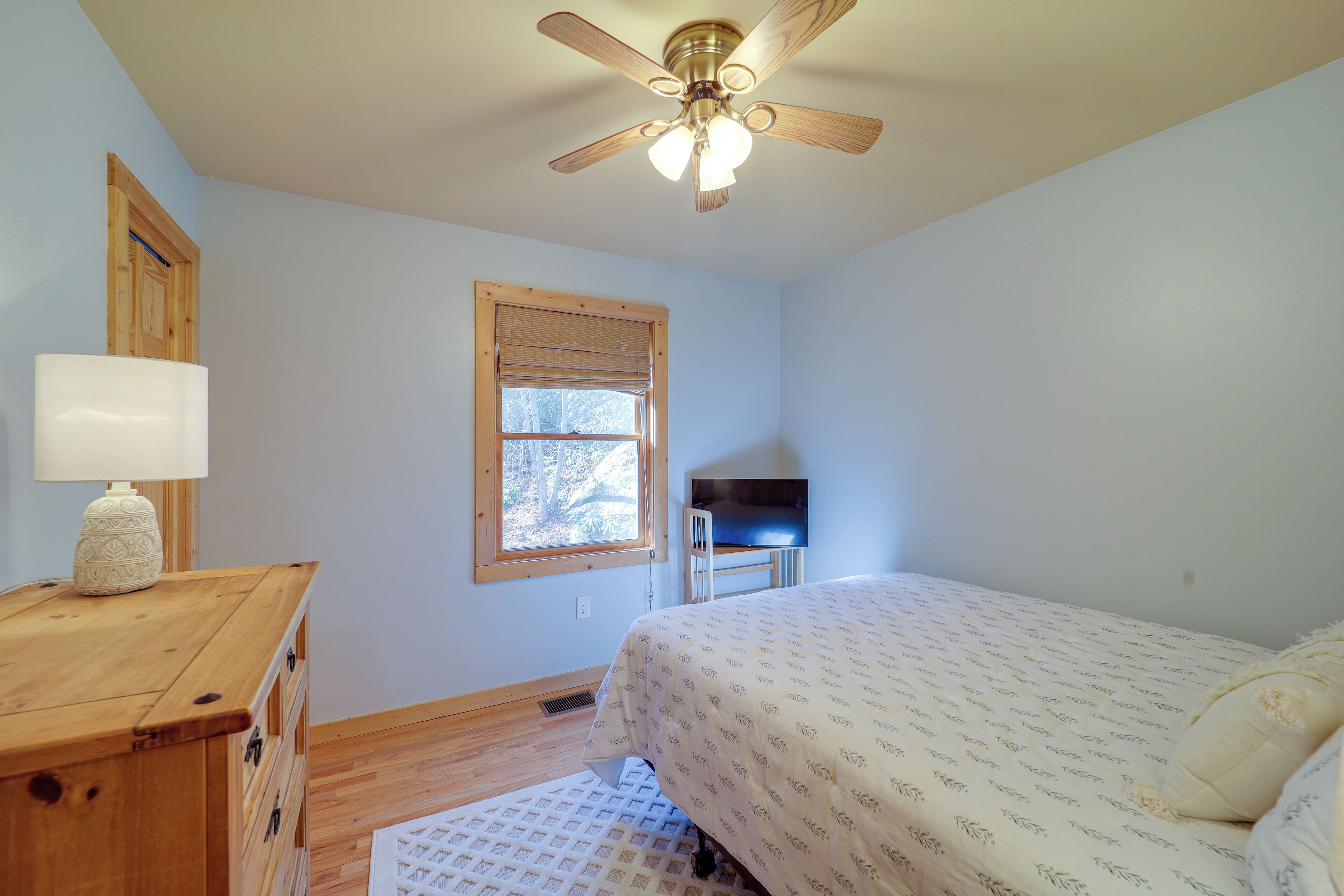 A bedroom with a bed, a wooden dresser, a ceiling fan, a window with blinds, and a television on a stand.