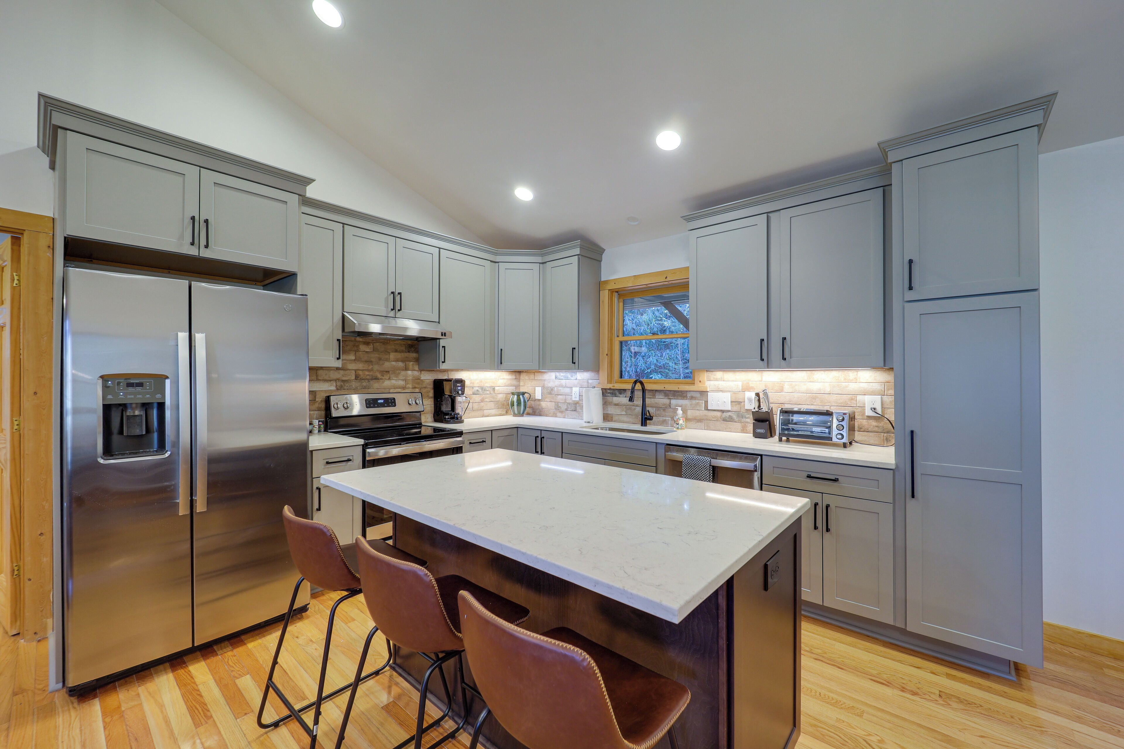 A modern kitchen with grey cabinets, stainless steel appliances, a central island with bar stools, and a window with a view.