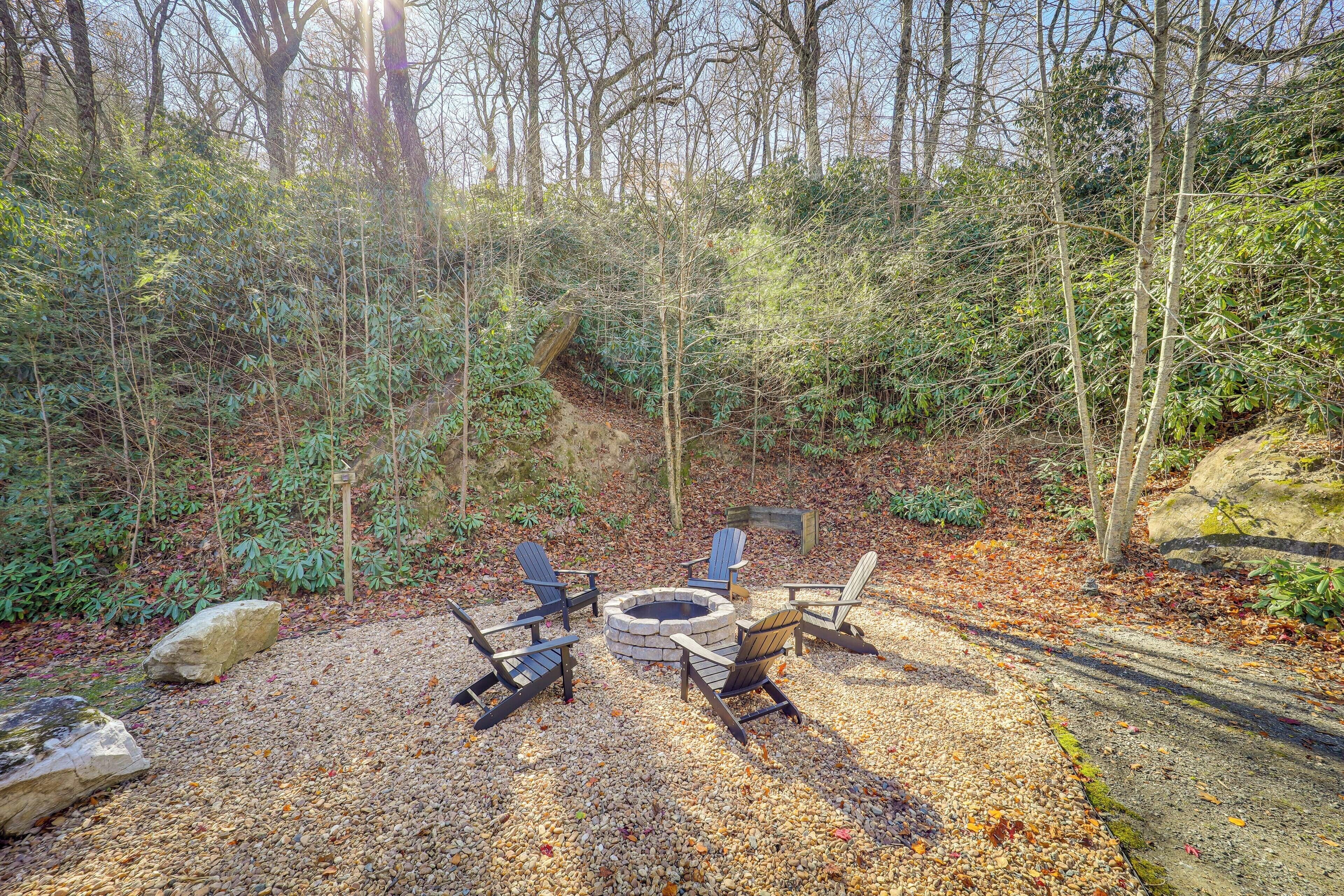 A forested area with a fire pit and four Adirondack chairs arranged around it.