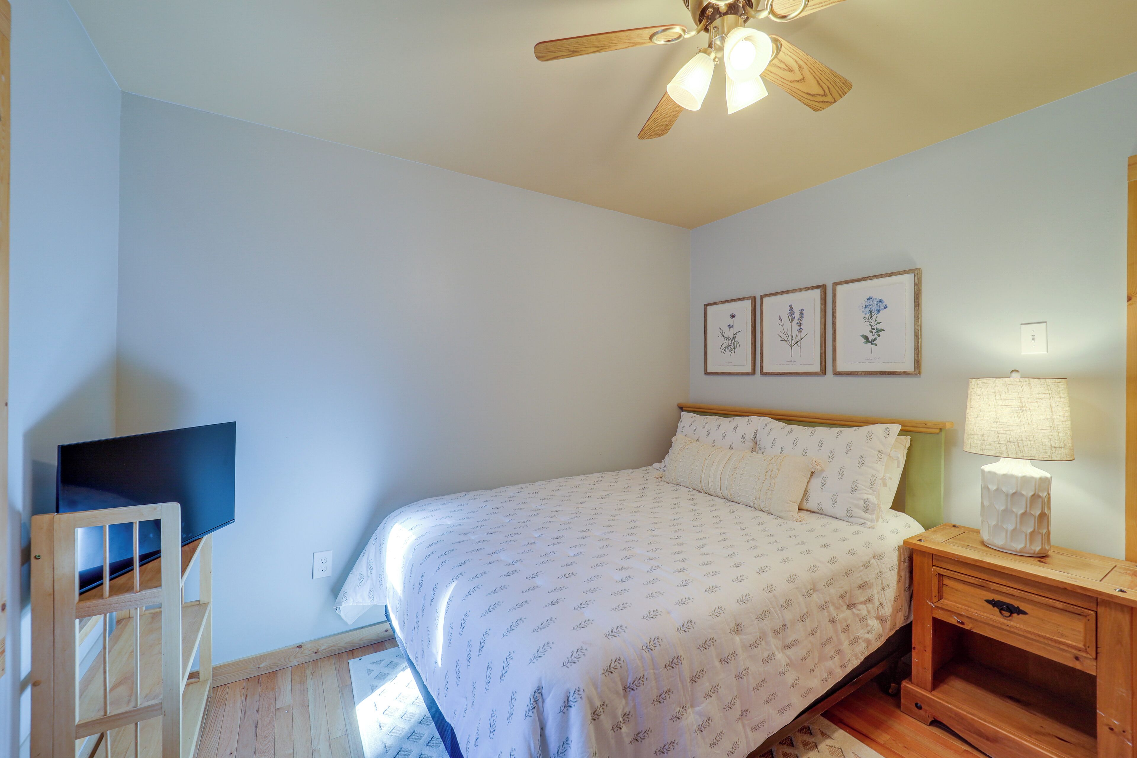 A bedroom with a bed, a wooden nightstand, a desk, a ceiling fan, and framed botanical prints on the wall.
