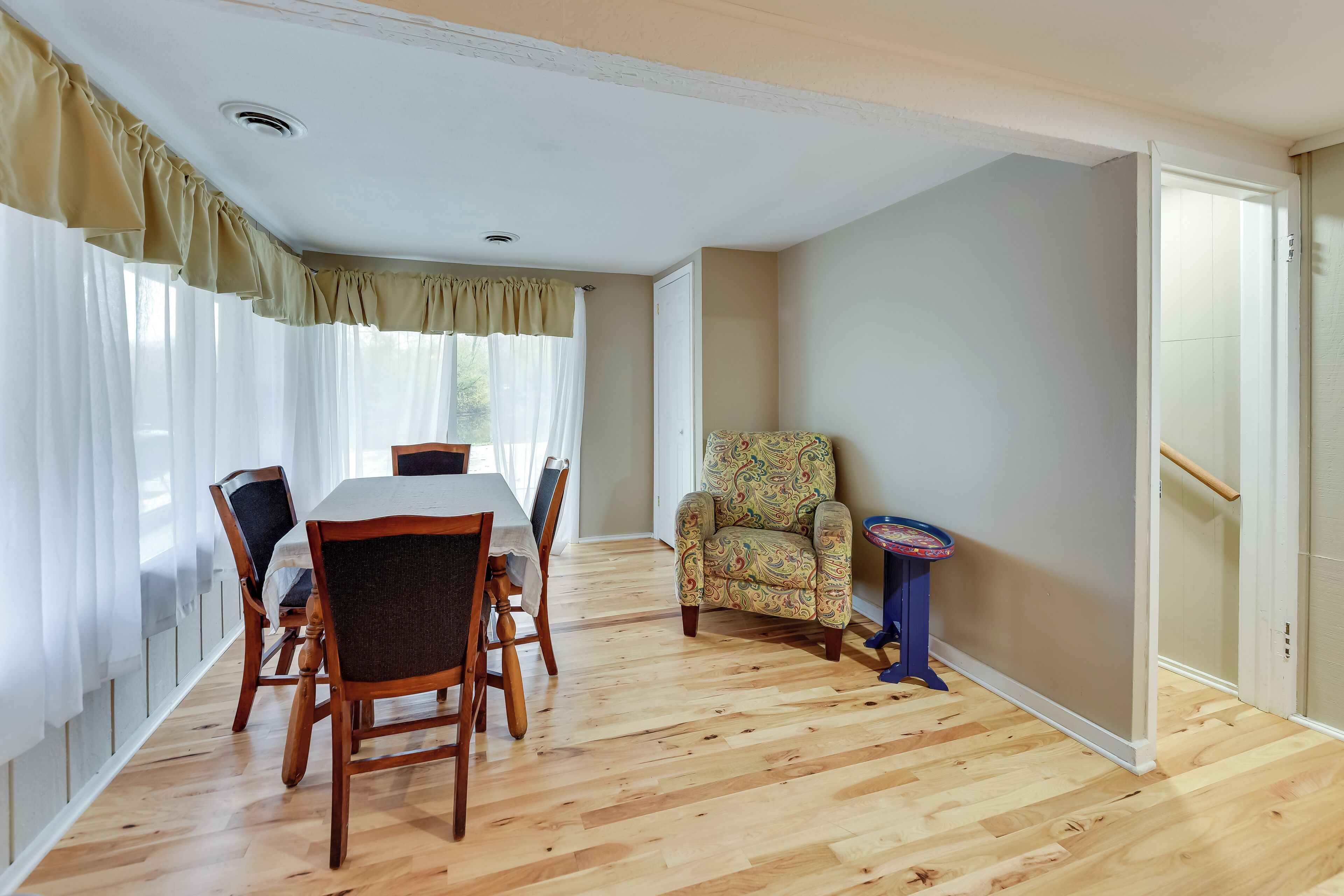 Un área de comedor con una mesa y sillas de madera, un sillón estampado y una mesita con cubierta colorida.