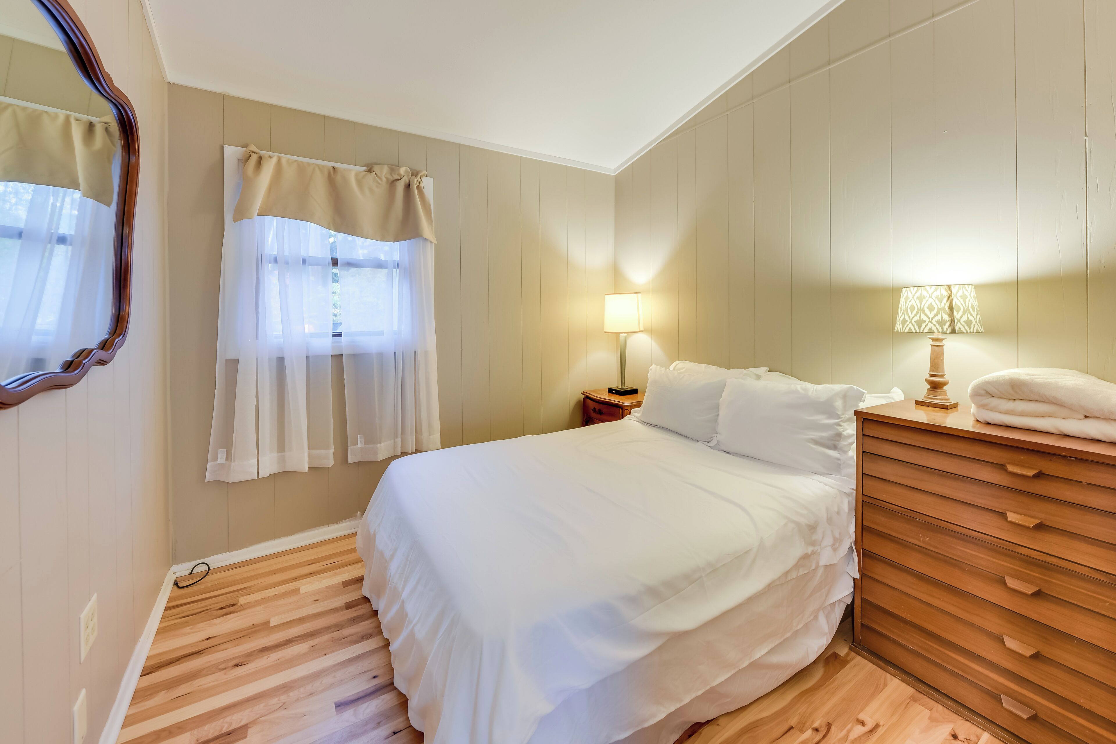 A bedroom with a bed, wooden dresser, two bedside lamps, a window with sheer curtains, and a mirror on the wall.
