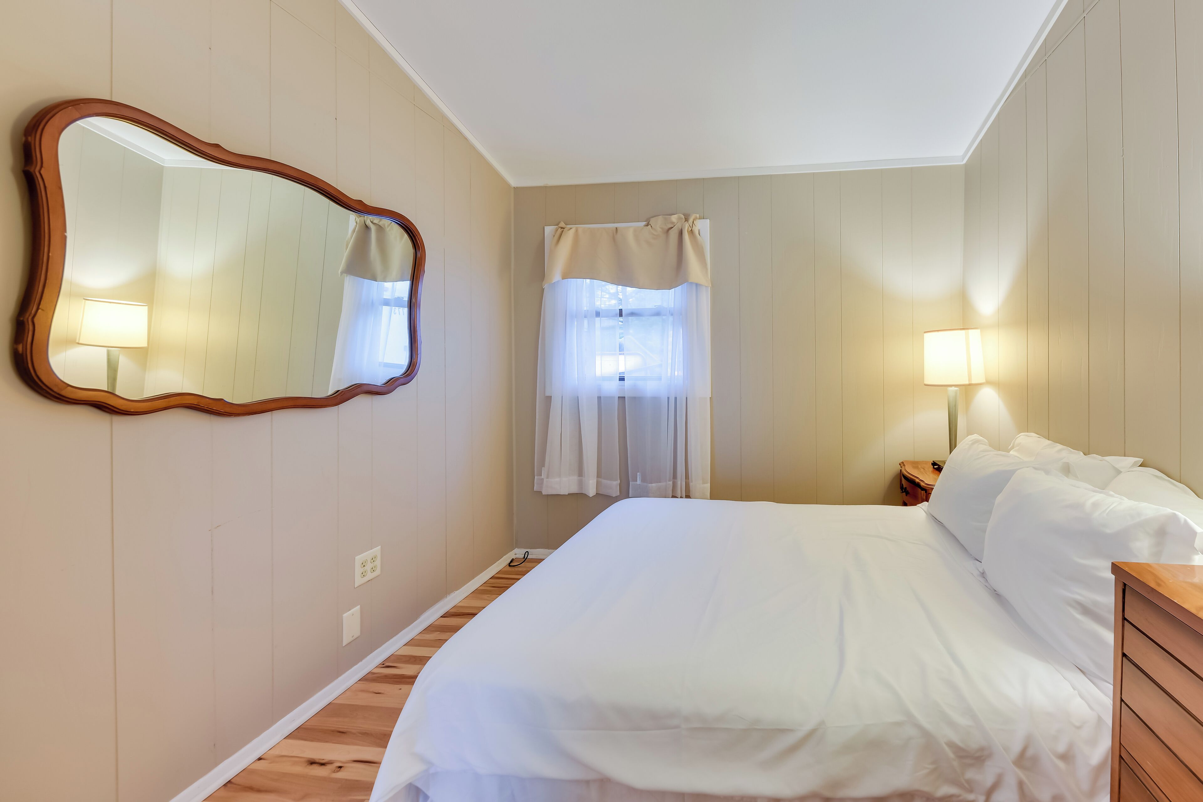 A bedroom with a large mirror, a bed with white linens, a nightstand with a lamp, and a window with sheer curtains.