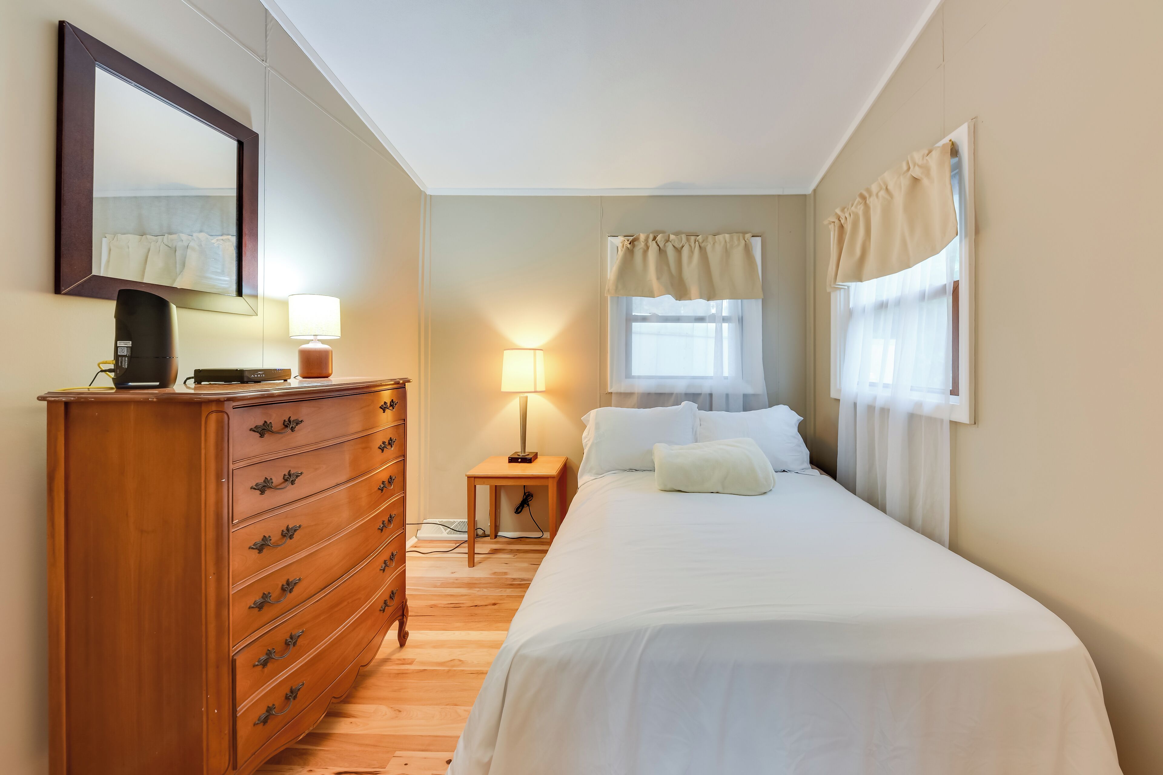 A bedroom with a wooden dresser, a bedside lamp, a bed with white linens, a window with curtains, and a mirror on the wall.