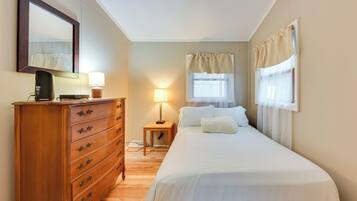 A bedroom with a wooden dresser, a bedside lamp, a bed with white linens, a window with curtains, and a mirror on the wall.