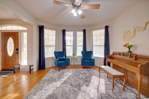 Un salón espacioso con piso de madera, ventilador de techo, un piano, dos sillones azules, un banco y un tapete de mármol.