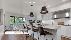 A modern kitchen with white cabinets, a large island with bar stools, a built-in oven, and a brick fireplace. - Expansive Monument Home: Hot Tub & Mountain Views! (Monument)