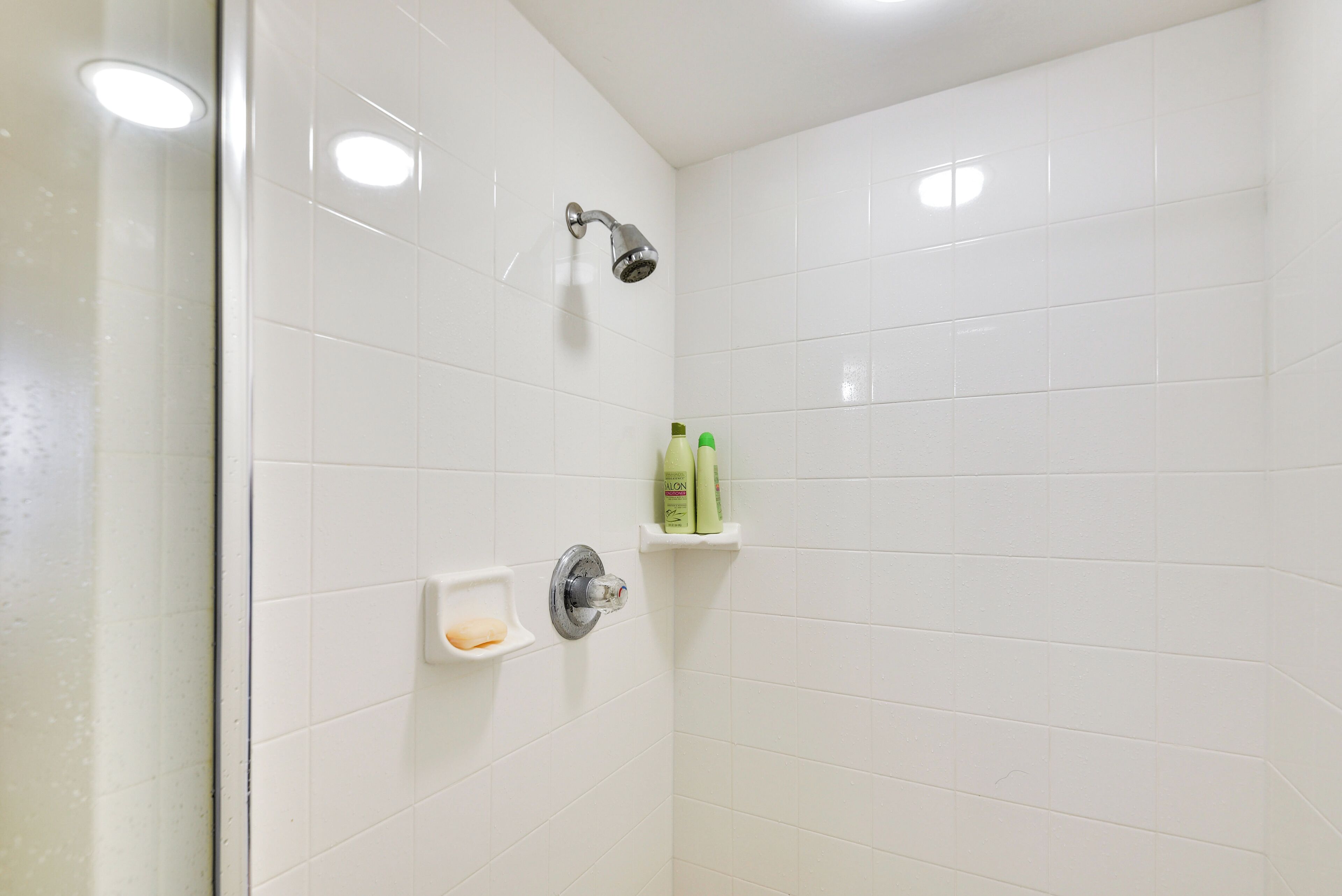 A bathroom with white tiles, a showerhead, and two green bottles on a shelf.