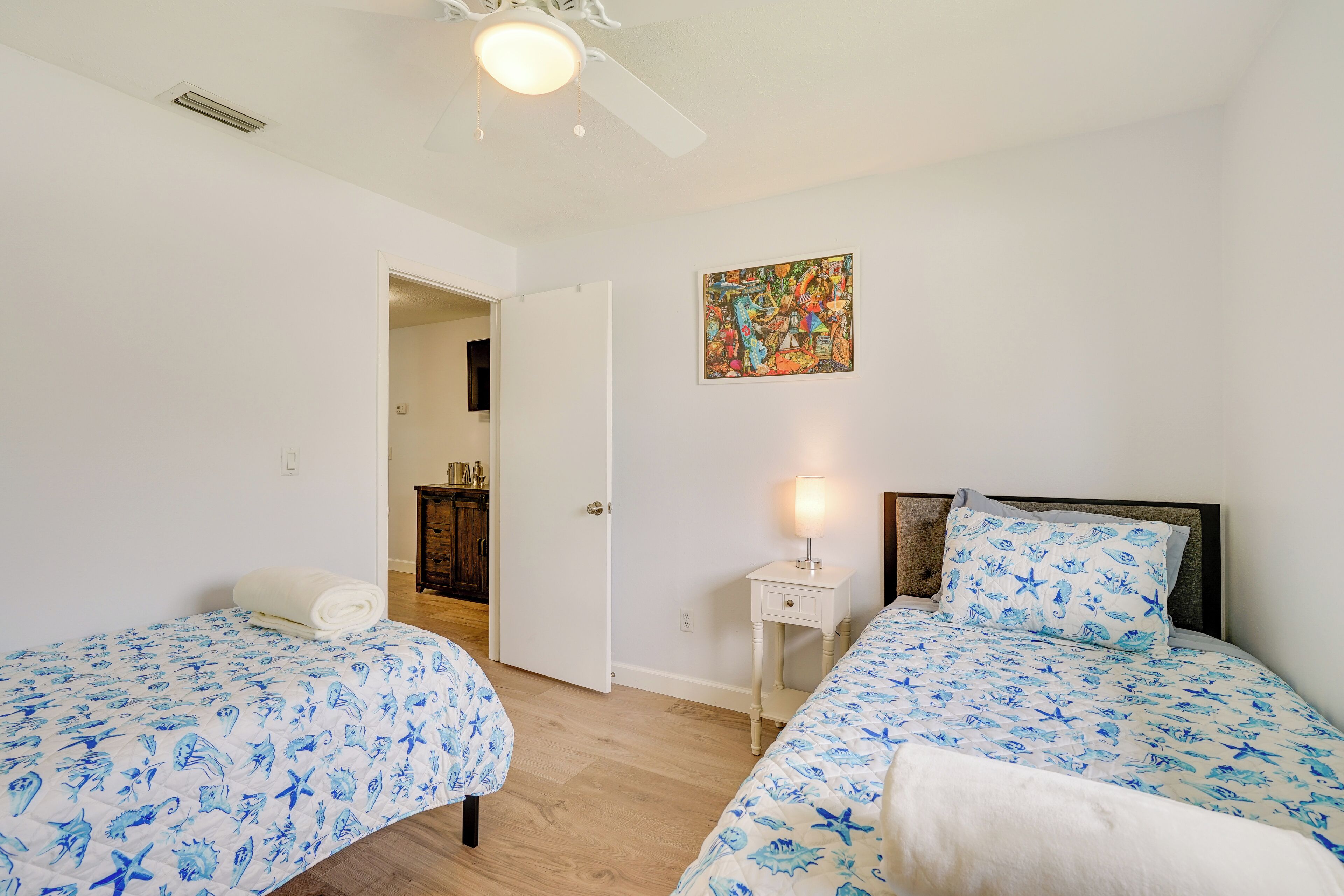A bedroom with two beds, a ceiling fan, a painting, and a bedside table with a lamp.
