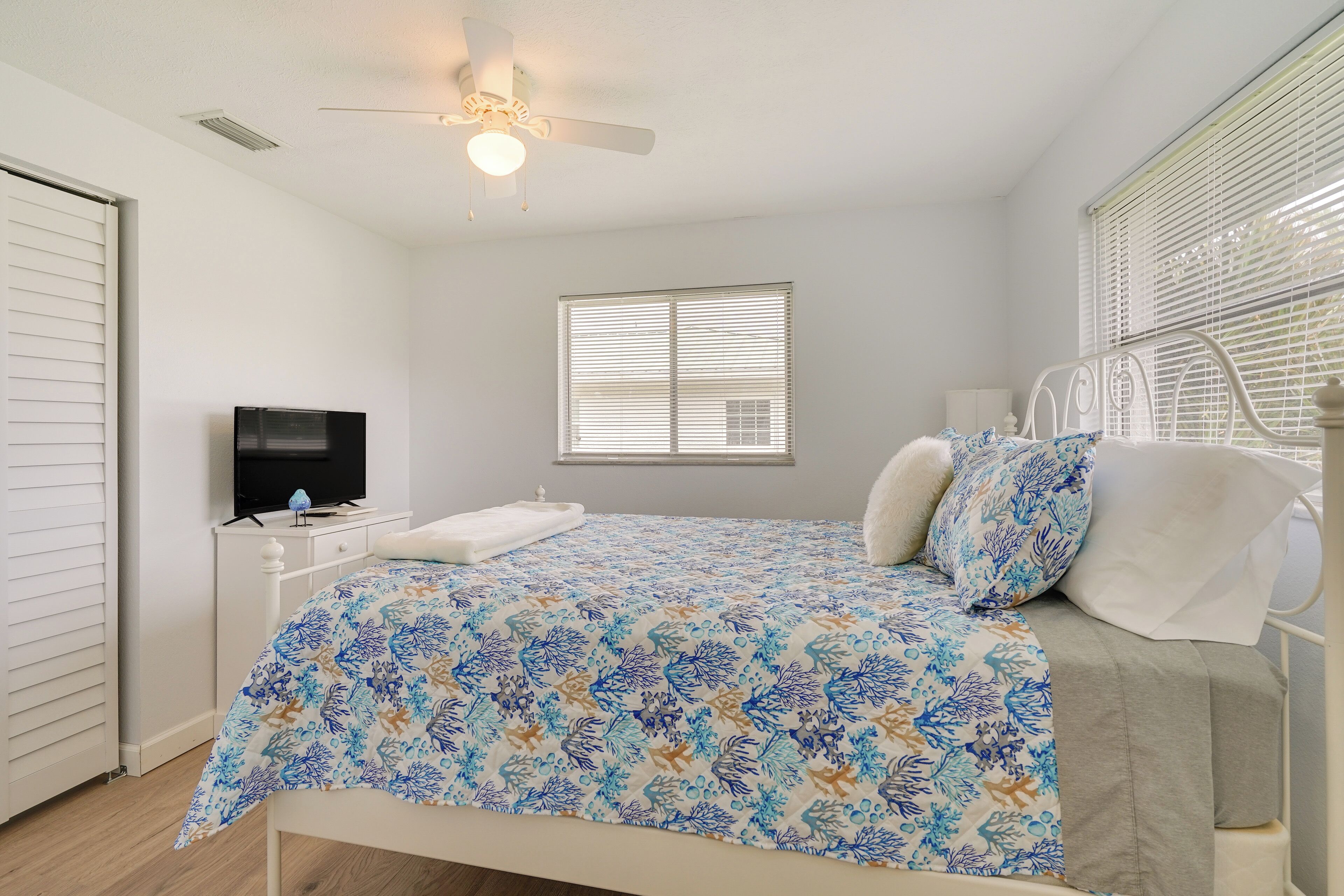 A bedroom with a bed, a ceiling fan, a television on a nightstand, and a window with blinds.