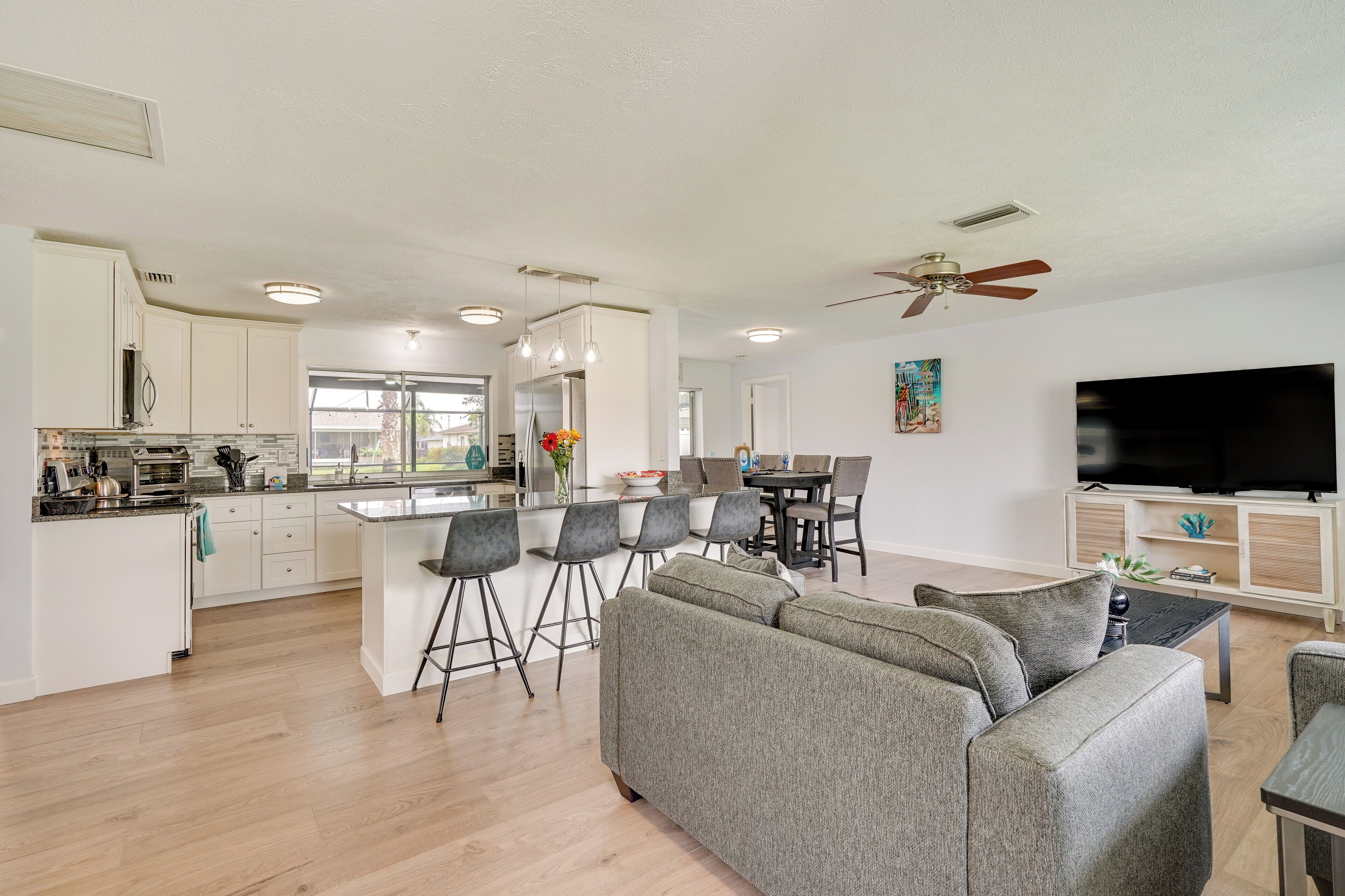 A modern kitchen with white cabinets, a dining area with a large TV, and a gray sofa in the living room.