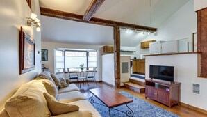 A living room with a sofa, coffee table, and TV stand. There is a dining area with chairs and a table. The room has wooden beams on the ceiling and a large window.