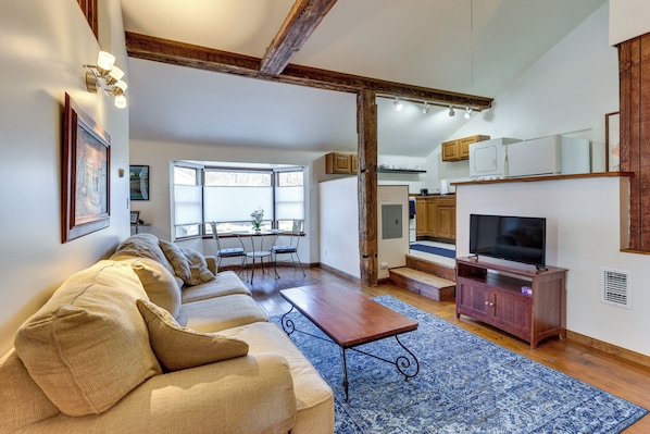 A living room with a sofa, coffee table, and TV stand. There is a dining area with chairs and a table. The room has wooden beams on the ceiling and a large window.