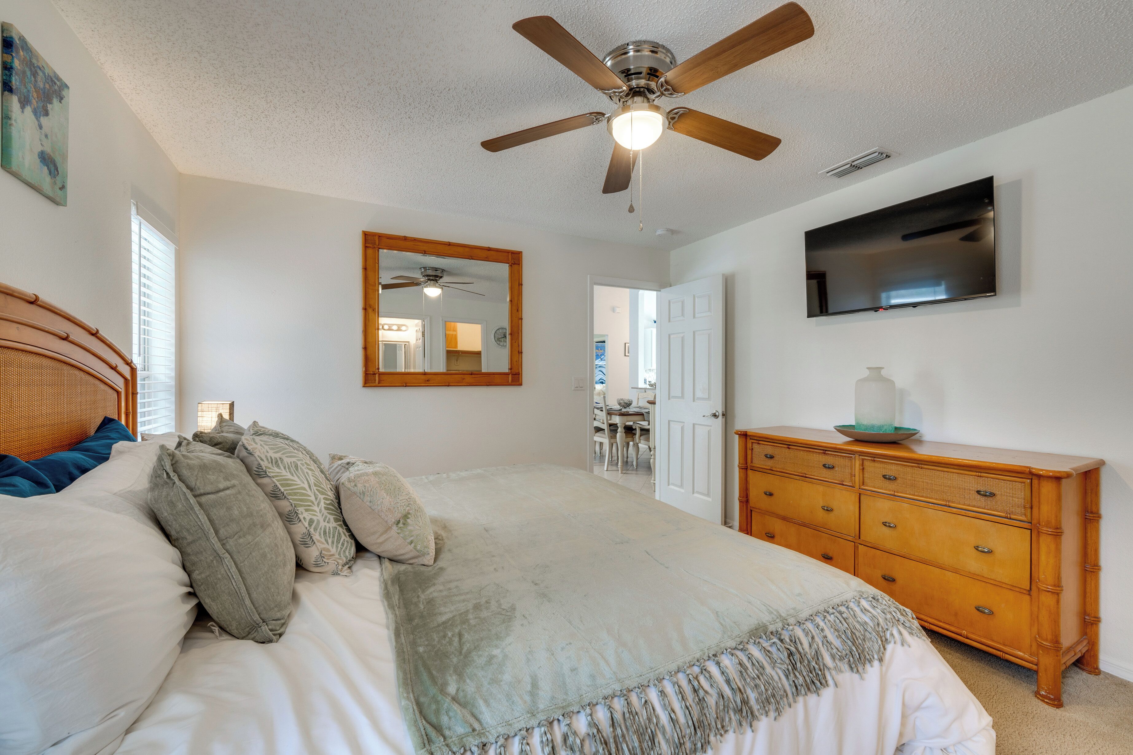 A bedroom with a bed, a wooden dresser, a ceiling fan, a flat-screen TV, and a mirror on the wall.