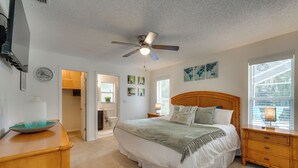 Une chambre à coucher comprenant un lit, une table de chevet en bois, un ventilateur de plafond, une fenêtre avec des stores et une porte donnant accès à une salle de bain.