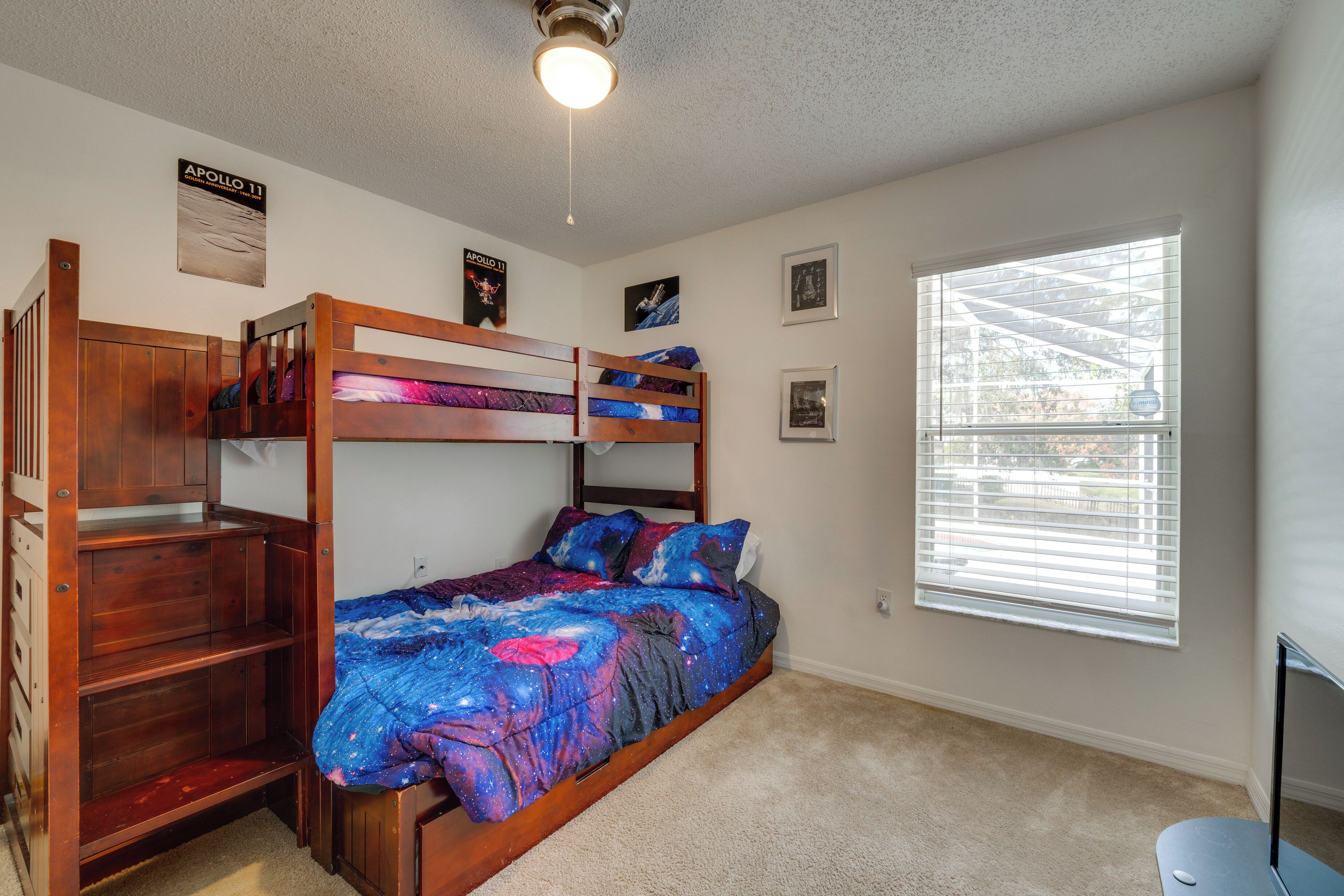 A bunk bed room with a galaxy-themed bedspread, a wooden desk, and a window with blinds.
