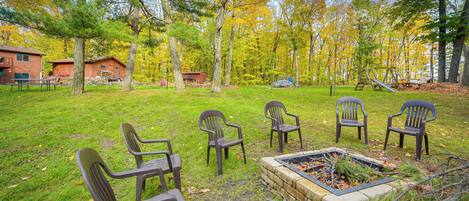 A grassy outdoor area with several Adirondack chairs, a fire pit, and a playground with a swing set.