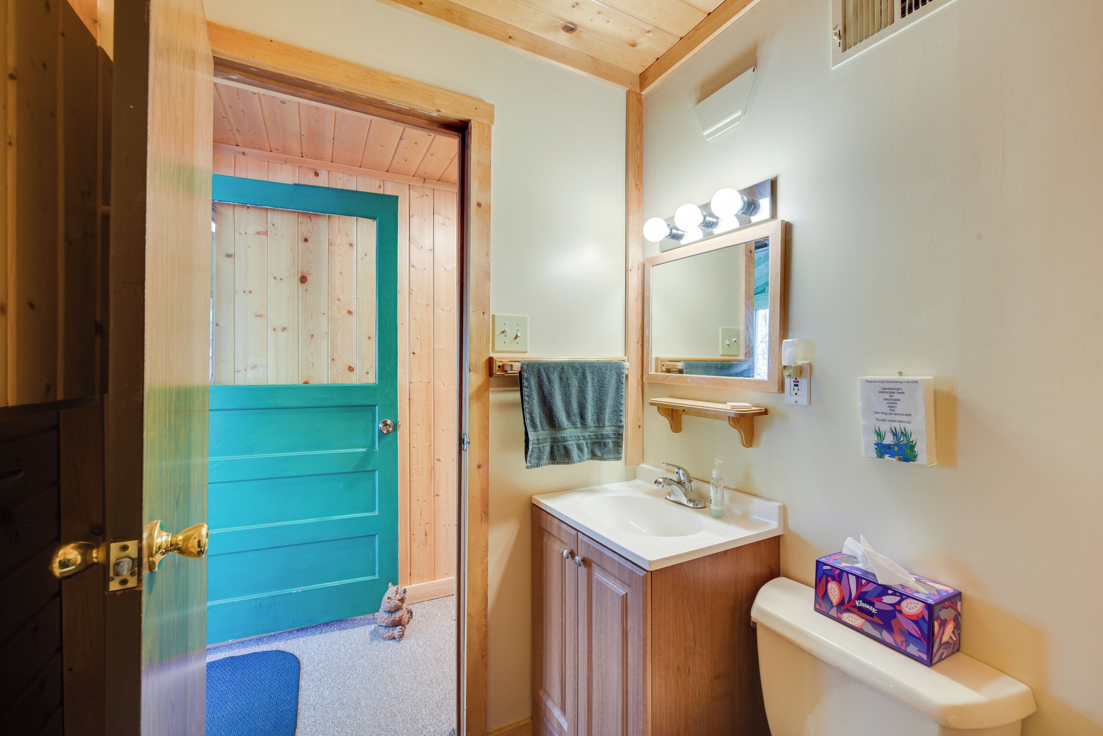 Un baño con puerta de madera, un lavabo, un espejo con luces y una caja de pañuelos sobre el inodoro.
