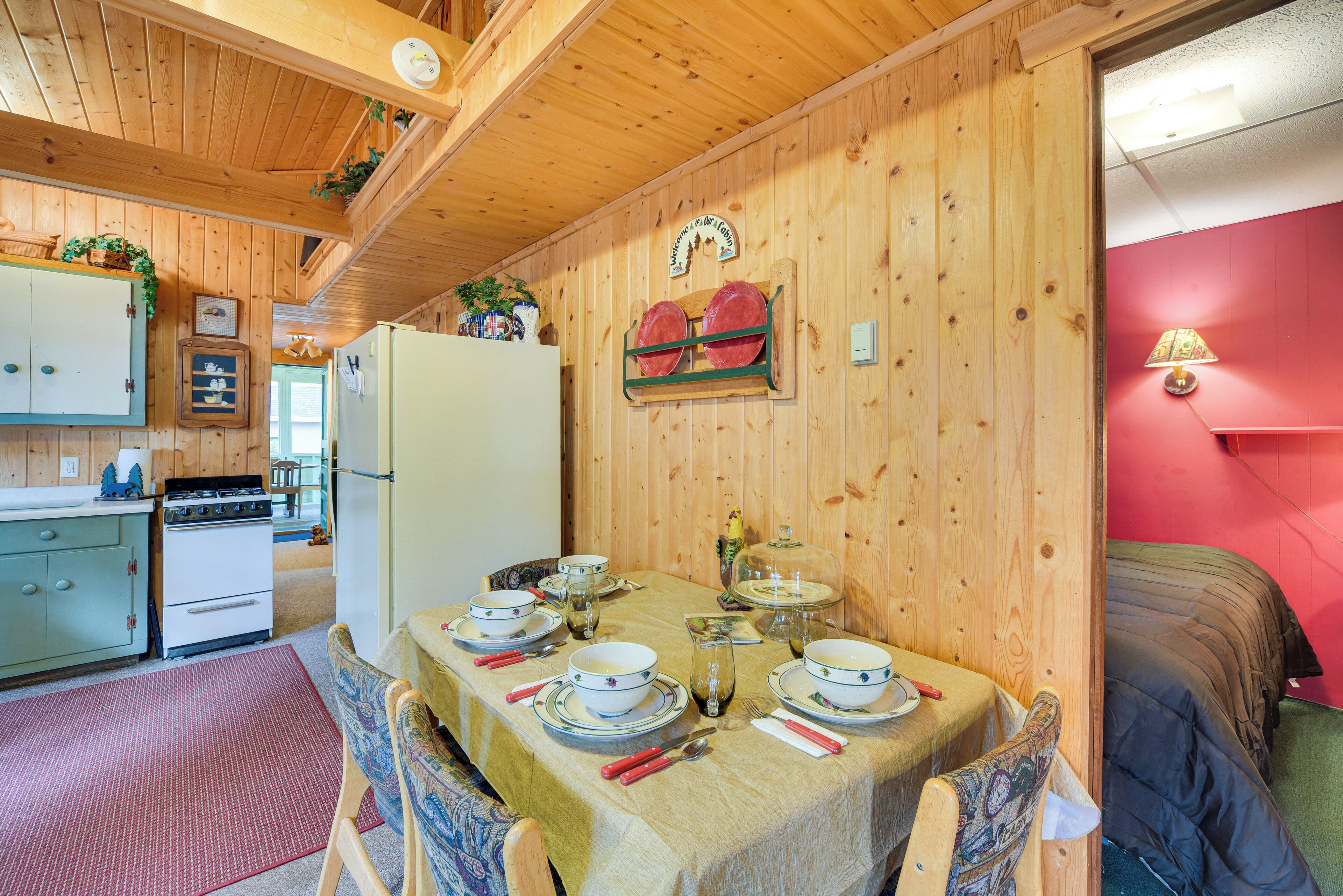 Interior de fusta amb taula de dinar per a quatre, cuina amb fogó i nevera, i paret vermella.