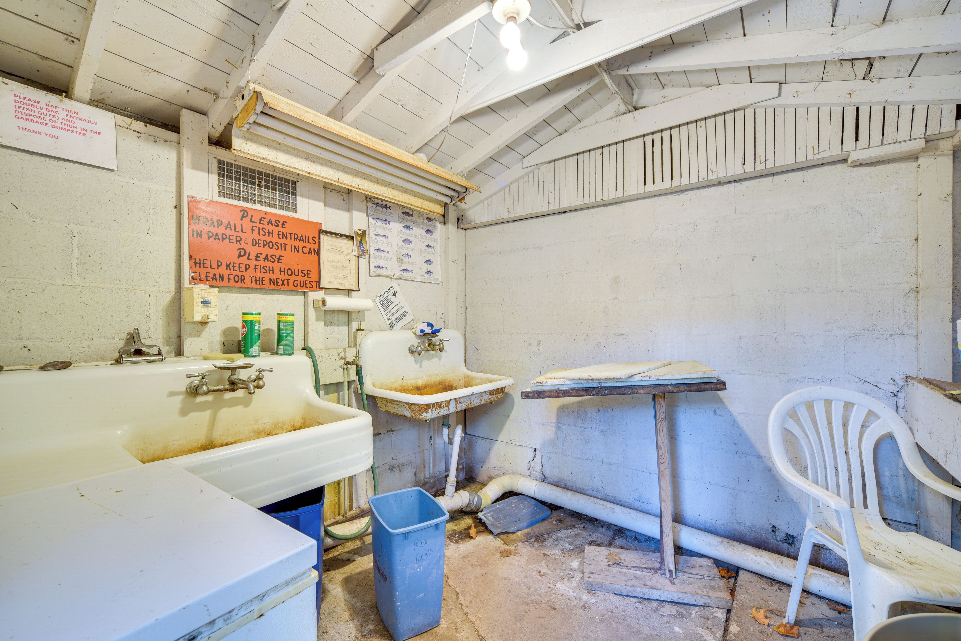A dilapidated bathroom with a large sink, a small window, and a sign requesting guests to wrap fish in paper and deposit it in the can.