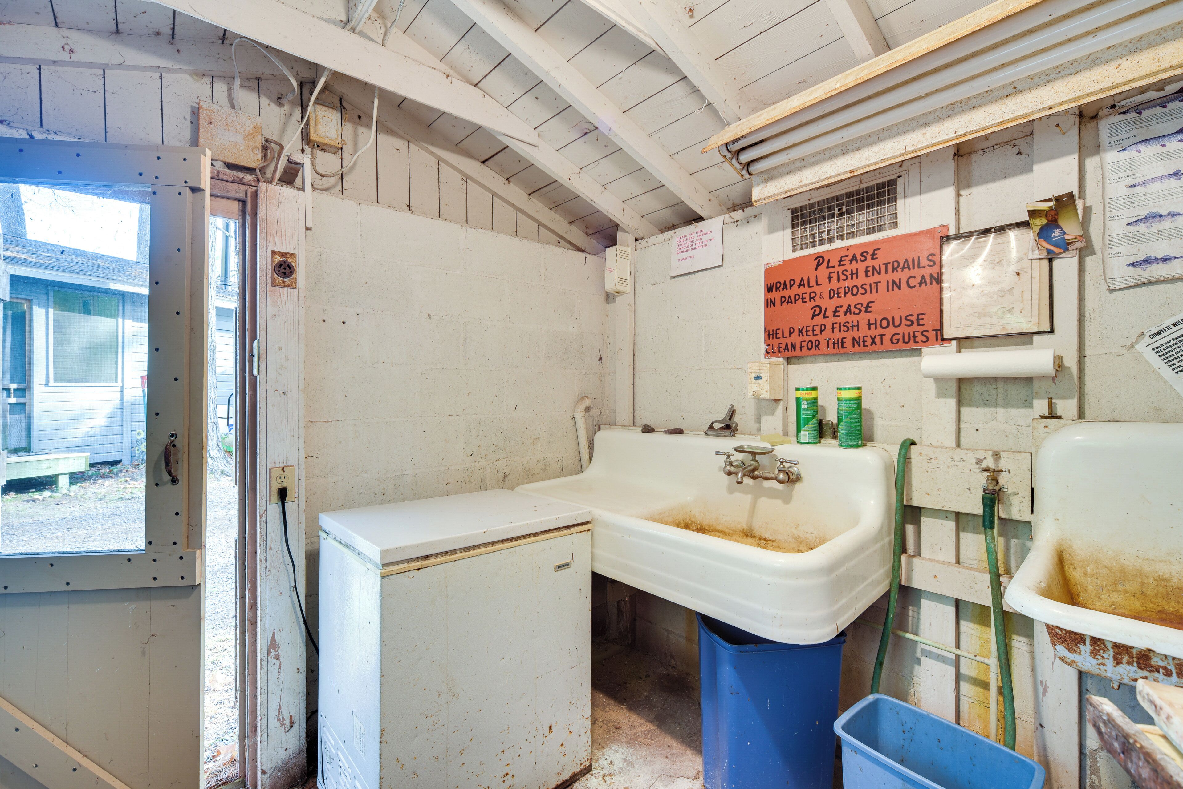 A dilapidated bathroom with a sink, a blue trash bin, and a sign requesting guests to wrap fish entrails and deposit them in a can.