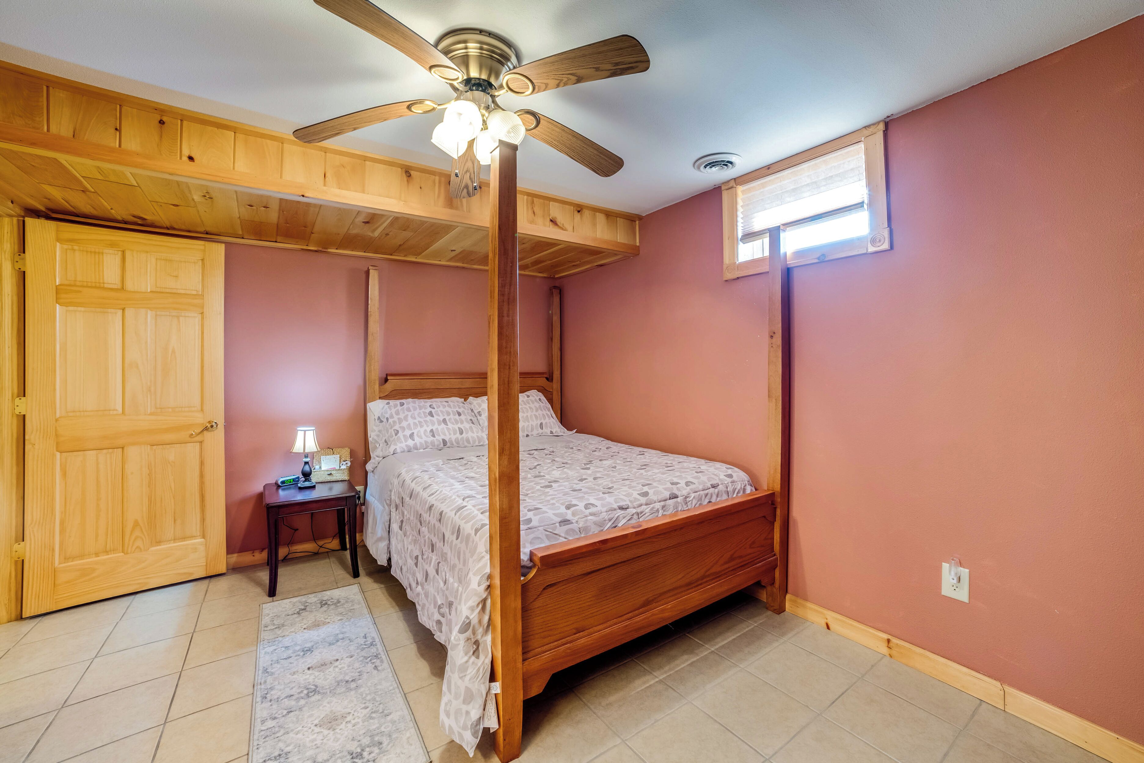 A bedroom with a four-poster bed, wooden ceiling beams, a ceiling fan, a small bedside table with a lamp, and a window with blinds.