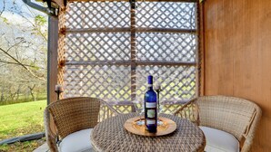 A wicker table with two chairs, a bottle of wine, and two glasses on a patio with a lattice screen in the background.
