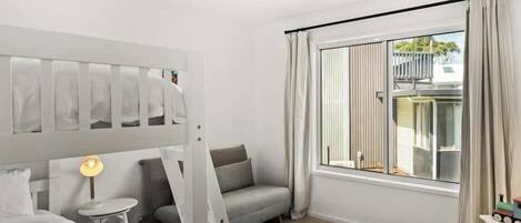 Um quarto com cama de solteiro, um sofá, uma mesinha redonda pequena, uma luminária, uma janela com cortinas e um pufe colorido.