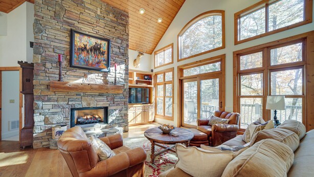 A spacious living room with a stone fireplace, leather furniture, and large windows offering a view of trees. - 5 Mi to Sapphire Valley Skiing: Serene Family Home (Sapphire)