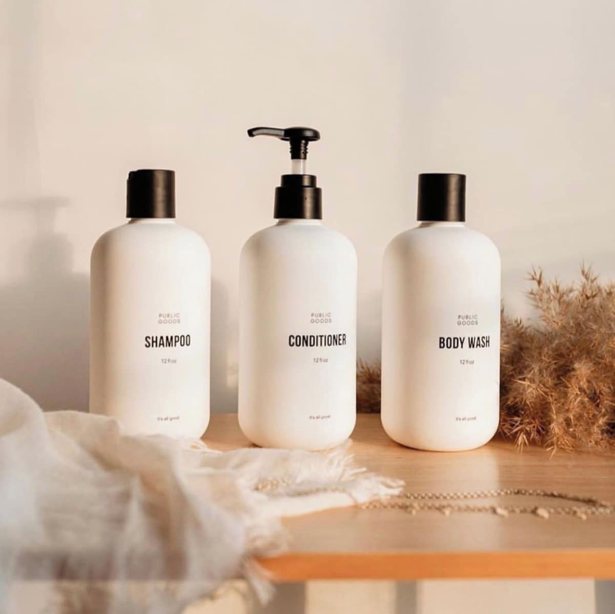 Drei weiße Flaschen mit Public Goods Produkten – Shampoo, Conditioner und Duschgel – jeweils mit Pumpspender, auf einer Holzoberfläche mit weißem Stoff und getrockneten Pflanzen im Hintergrund.