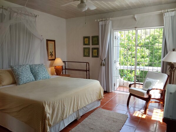 A bedroom with a bed, a chair, a lamp, and a view of greenery through a window. - Large Secure 2 Bedroom Apartment with large pool steps to Mullins Beach (St Peter)