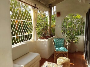 A sunlit patio with a white cushioned bench, a wicker chair with a patterned cushion, and a woven pouf. - Large Secure 2 Bedroom Apartment with large pool steps to Mullins Beach (St Peter)