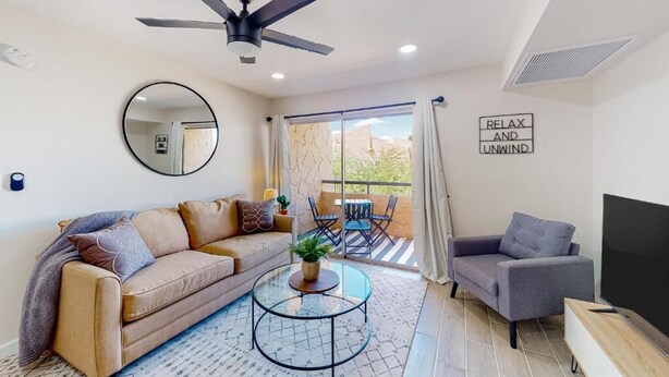 Ein modernes Wohnzimmer mit Sofa, Sessel, Couchtisch und Fernseher. Es gibt einen Deckenventilator, einen großen Spiegel und einen Balkon mit Blick.