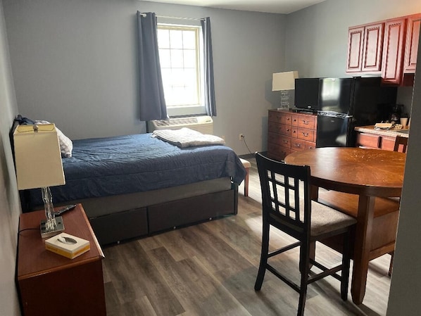 A compact room with a bed, a desk, a chair, a small kitchenette, and a window with curtains.