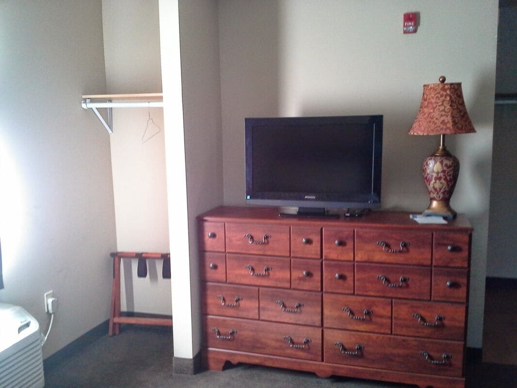Une commode en bois, un téléviseur à écran plat et une lampe, un porte-manteau et un détecteur d’incendie fixés au mur.