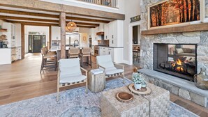 A spacious living area with a stone fireplace, wooden beams, and a view of the kitchen. - Custom built Adirondack style home sleeps 16 in 6 bedrooms (Moultonborough)