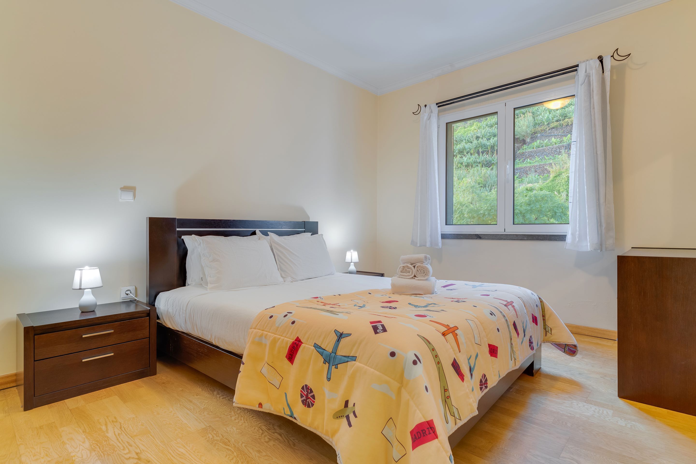 Une chambre à coucher avec un lit, des tables de chevet, une fenêtre avec des rideaux et une vue sur la verdure à l’extérieur.