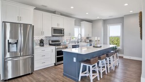 A modern kitchen with white cabinets, stainless steel appliances, a central island with bar stools, and a view of greenery outside. - Luxury Coastal Escape | Heated Pool | Golf Cart Included (Gulf Shores)