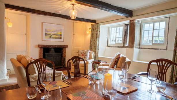 A dining room with a wooden table set for four, surrounded by chairs, a fireplace, and a window with curtains. - Townend Farmhouse - Watermillock, Ullswater (Watermillock)