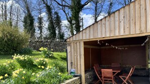 A wooden building with a stone wall, a garden with daffodils, and a table with chairs. - Townend Farmhouse - Watermillock, Ullswater (Watermillock)