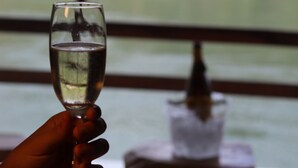 A hand holding a glass of white wine, with a view of a river and buildings in the background.
