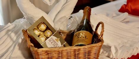 A wicker basket containing chocolates, a bottle of wine, and a bottle of champagne, placed on a red bedspread with heart-shaped towels.