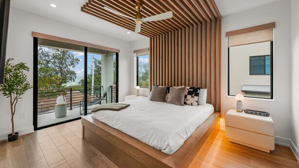 A modern bedroom with a large bed, wooden ceiling slats, a ceiling fan, a bedside table with a lamp, and a view of the outdoors through large windows. - Brand New home with Incredible Rooftop Deck and Pool! - Just Breathe (Anna Maria)