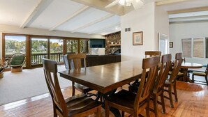 A spacious dining area with a large wooden table and chairs, a ceiling fan, and a view of a house through large windows. - The Lake House on Powers |Grassy Frontage |Lakeside Deck |Rec Room w/Pool Table & Bar |Fire Pit (Genoa City)