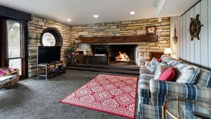A cozy living room with a stone fireplace, a television, and a plaid sofa. - The Lake House on Powers |Grassy Frontage |Lakeside Deck |Rec Room w/Pool Table & Bar |Fire Pit (Genoa City)
