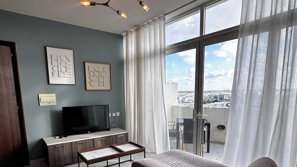 A modern bedroom with a large bed, a TV, a desk, and a view of the city through large windows. - Central & Spacious Penthouse w/ 1BR + City View (L-Imsida)