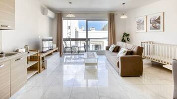 Un appartement moderne comprenant une cuisine, un coin repas et un salon. Il dispose d’une grande fenêtre offrant une vue sur les immeubles et d’un balcon.